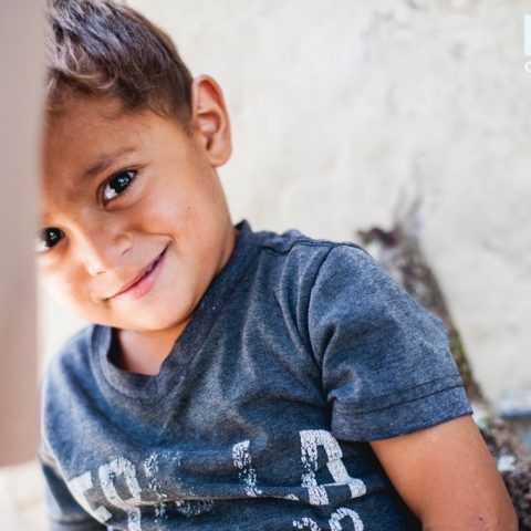 A playful 3-year-old boy smiles and hides behind his mom as she talks about the many needs her family faces. His family needs a new roof on their mudbrick home.